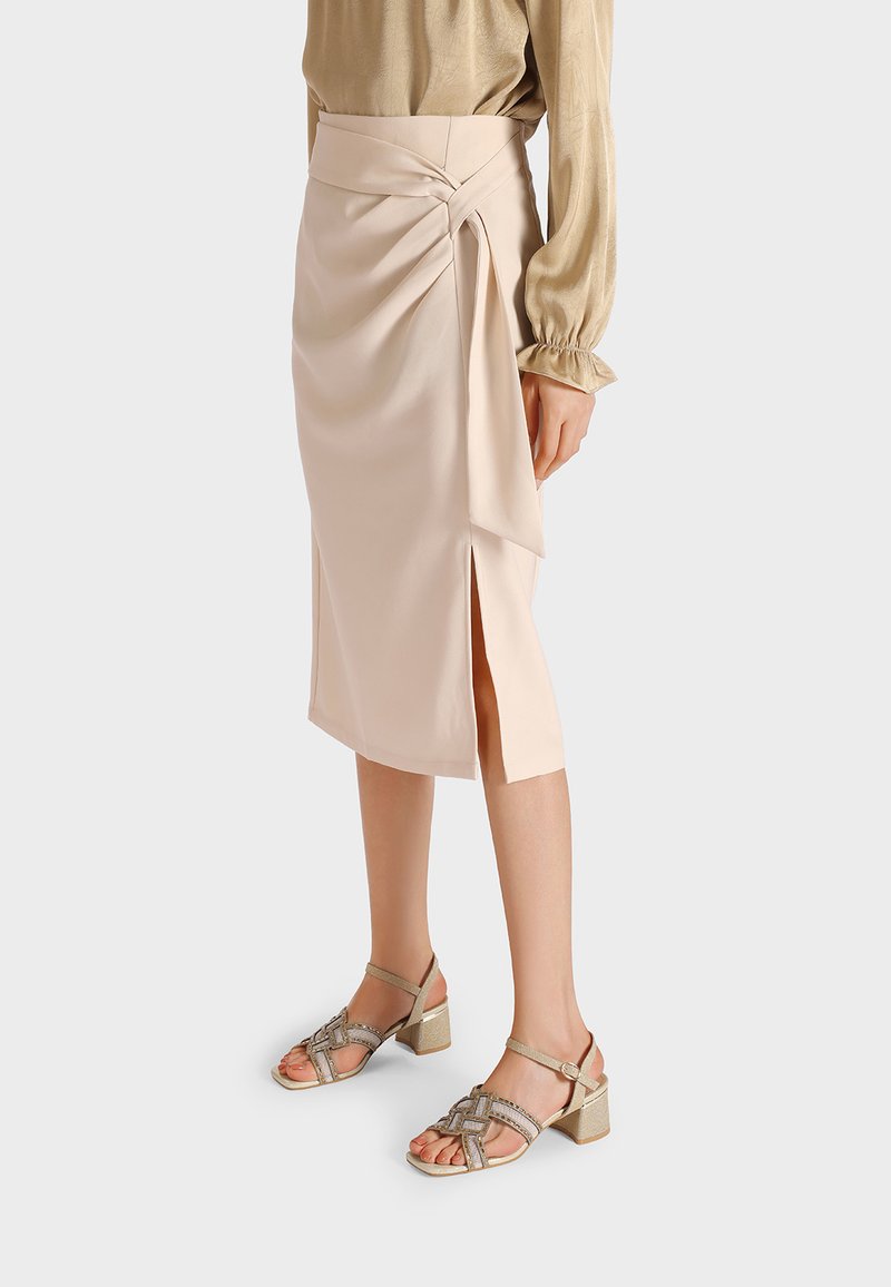 Gonna beige a lunghezza ginocchio con design a portafoglio, caratterizzata da uno spacco laterale e una texture liscia. Abbinata a sandali con tacco a blocco metallici.