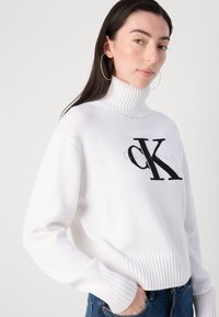 Hvitstrikket genser med høy ribbestrikket krage, med en stor svart "CK"-logo. Ribbestrikkede mansjetter og nederkant gir tekstur og struktur.