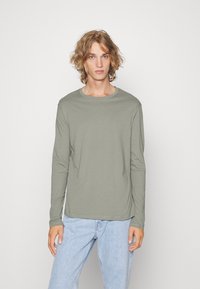 Langermet skjorte i lysegrønn, laget av mykt stoff. Har rund hals og avslappet passform, kombinert med lyseblå jeans.