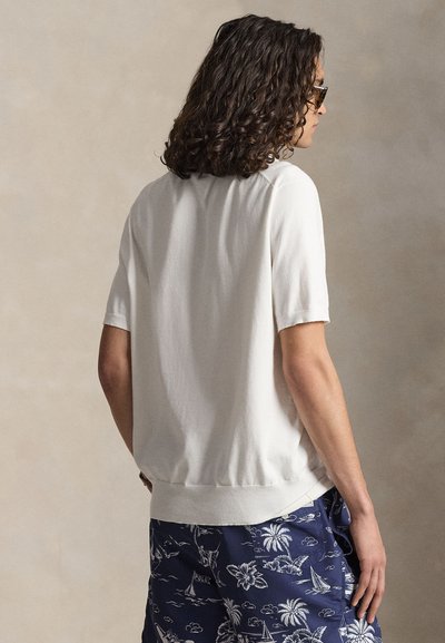 Άτομο με σγουρά μαλλιά που φοράει γυαλιά ηλίου, λευκό κοντομάνικο πουκάμισο και σκούρες μπλε βερμούδες με τροπικό σχέδιο, σε πλάγια όψη με ουδέτερο φόντο.