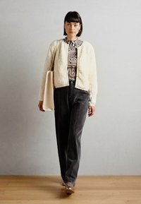 Cream faux fur bomber jacket over a black and white patterned top, paired with gray trousers and a beige bag, set against a neutral backdrop.
