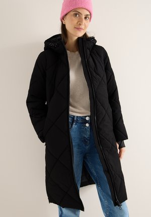 Sort quiltet lang frakke med lynlås, hætte og sidelommer, båret over en beige top og blå jeans, mod en neutral baggrund.