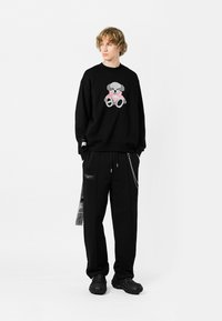 Sudadera negra con un gráfico de un oso, combinada con pantalones negros holgados que tienen un bolsillo lateral y detalles de cadena. El modelo lleva zapatillas negras gruesas.