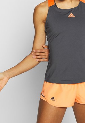 Osoba ubrana w czarny i pomarańczowy bezrękawnik Adidas oraz pomarańczowe spodenki Adidas wyciągająca rękę przez tors.
