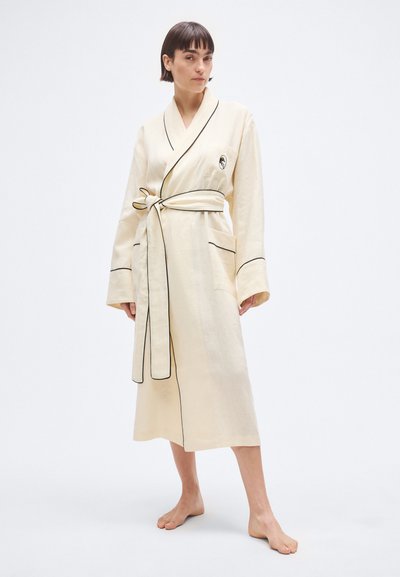 Bēša halāts ar melnām apmalēm, garām piedurknēm un jostas auklu jostasvietā. Aprīkots ar divām sānu kabatām un logotipu krūtīs. Garums līdz vidus teļiem.