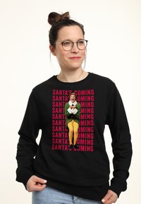 Felpa nera con un grafico centrale di un personaggio in un vestito verde con gambe gialle, circondato da testo ripetuto: "STA ARRIVANDO SANTA".