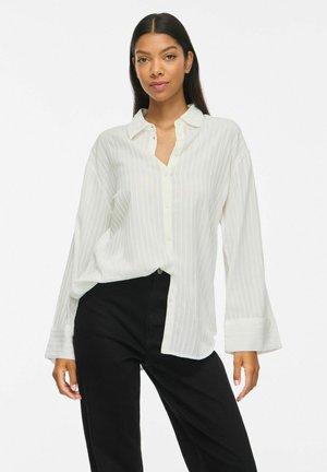 Femme aux longs cheveux foncés portant une chemise blanche rayée ample à boutons et un pantalon noir sur un fond clair uni.