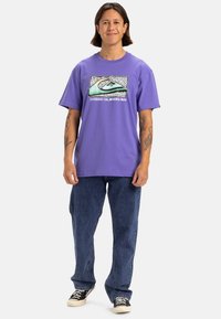 Camiseta de algodón morada con un estampado gráfico que presenta una ola, combinada con unos vaqueros holgados de mezclilla y zapatillas de lona negras.