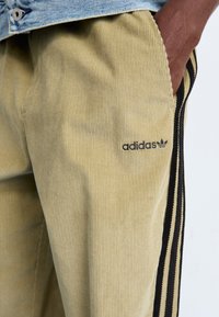 Buks i lyse beige fløjl med lodrette ribber, prydet med et sort Adidas-logo og sidesømme med sort og hvid striber.