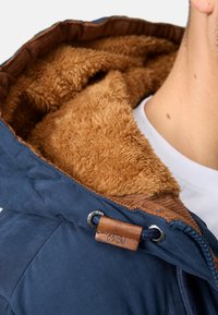 Veste à capuche bleu marine avec des finitions en velours côtelé marron et une doublure en polaire douce couleur beige. Comprend une étiquette en cuir et un système de cordon de serrage.
