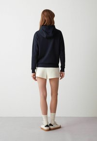 Mörkblå huva-tröja med avslappnad passform, kombinerad med ljusa shorts. Modellen visar ryggen för att framhäva materialets textur och enkla design.