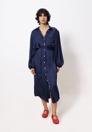 Robes chemises en satin bleu marine avec manches longues, boutonnage devant et taille cintrée par une ceinture. Associe à des ballerines rouges vives avec un design découpé.