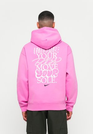 Person i en lys pink hoodie med hvid grafisk tekst "Bevæg din krop, bevæg din sjæl" og et sort Nike-logo på ryggen, vendt væk.