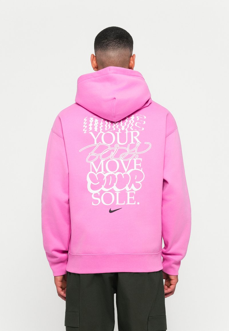 Asmuo, vilkintis ryškiai rožine džinsine striuke su balta grafine užrašu "Judėk savo kūną, judėk savo sielą" ir juodu "Nike" logotipu ant nugaros, atsukęs veidą atgal.