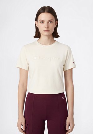 Giovane donna con capelli castani lisci, indossa una maglietta Champion a maniche corte color crema e pantaloni color bordeaux, in piedi davanti a uno sfondo bianco.