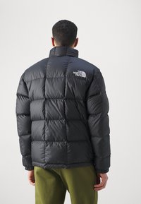 Man i svart pufferjacka med The North Face-logotyp på ryggen, i kombination med olivgröna byxor, vänd bort mot en neutral bakgrund.