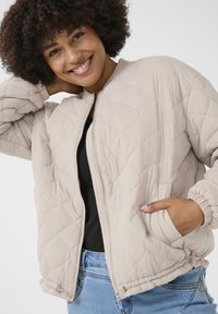Veste bomber matelassée beige avec fermeture éclair, poignets côtelés et poches latérales. Texture douce, tissu léger et design décontracté.