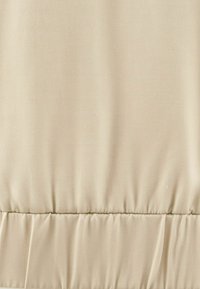 Tessuto beige con una texture liscia, caratterizzato da dettagli arricciati lungo la cucitura inferiore. La superficie ha una leggera lucentezza.