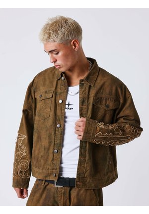Jeune homme aux cheveux blonds courts portant une veste marron avec des manches brodées sur une chemise blanche, regardant vers le bas et tenant le devant de la veste.