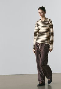 Giovane donna che cammina al chiuso indossando una maglia a maniche lunghe beige e nera a righe, pantaloni marroni larghi con coulisse, e scarpe nere.