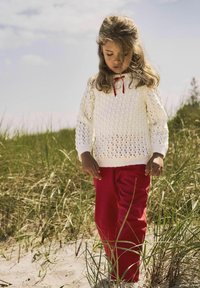 Vit stickad tröja med en strukturerad väv och röd bandaccent, kombinerad med klarröda byxor. Mot en bakgrund av gräs och sand.