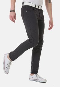 Schwarze, strukturierte Slim-Fit-Jeans mit einem weißen Gürtel, kombiniert mit weißen Turnschuhen, die rote und blaue Akzente aufweisen, in einer stehenden Pose präsentiert.