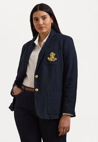 Marineblazer i en strukturert design, med gullknapper og en brodert lomme med logo. Sammen med en hvit skjorte og mørke jeans.