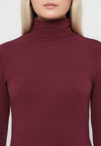 Burgundy turtleneck-paita, joka on valmistettu pehmeästä ja joustavasta kankaasta. Ominaisuuksiin kuuluu tiimalasimuotoilu, korkea kaulus ja pitkät hihat, sekä sileä pinta.