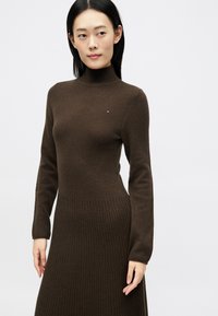 Γυναίκα με ίσια μαύρα μαλλιά, που φοράει ένα εφαρμοστό σκούρο καφέ ριμπ τζιν φόρεμα με ζιβάγκο σε λευκό φόντο.