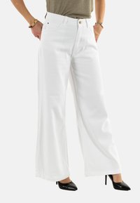 Pantalones de mezclilla anchos y blancos, con botón y presillas para cinturón, combinados con zapatos de tacón negro. Textura suave y ajuste hecho a medida.