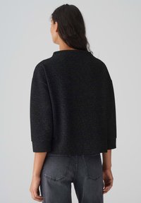 Schwarze, strukturierte Jacke mit hohem Kragen und Dreiviertelärmeln. Getragen über grauen Jeans mit boxy Schnitt und minimalistischem Design.