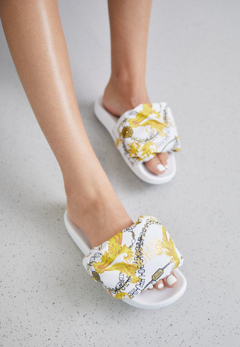 Witte schuif sandalen hebben een dikke band versierd met een geel en gouden barokpatroon, inclusief kettingen, en een glanzende afwerking.