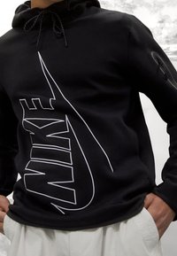 Sudadera negra con capucha que presenta un gran logo de Nike en blanco en la parte frontal, con un tejido texturizado y cordones ajustables en la capucha.
