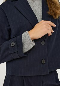 Námořnický pruhovaný sáčko s cropped designem, spárované se šedým pleteným svetrem. Viditelné černé knoflíky na rukávech a límci.