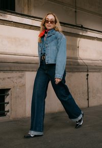 Femeie care merg pe jos în aer liber, purtând o jachetă din denim, blugi evazați, un fular roșu la gât, ochelari de soare și pantofi alb-negru pe o stradă pavata.