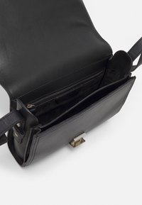 Černá kožená crossbody taška s hladkým povrchem, která má zipovou kapsu, přední klopu a kovové zapínání. Uvnitř jsou různé přihrádky.