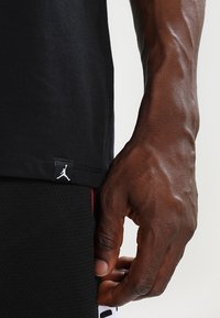Svart bomullströja med en liten vit Jumpman-logotyp vid fållen, parat med strukturerade svarta shorts med röda detaljer.
