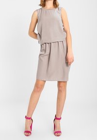 Ärmelloses, hellgraues Kleid mit gerafftem Bund und fließendem Oberteil. Kombiniert mit leuchtend roten High-Heels mit Knöchelriemen.