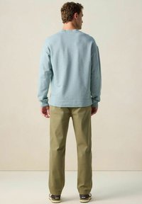 Homme portant un sweat-shirt bleu clair, un pantalon vert olive et des baskets noires et blanches, se tenant face à un fond beige uni.