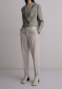 Olivově zelený vypasovaný dvojitý sak přes béžové kalhoty s vysokým pasem, doplněné zelenými podpatkovými kotníkovými botami a odpovídajícími pletenými ponožkami.