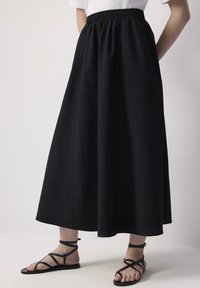 Jupe noire, longueur cheville, en tissu doux avec une taille élastique ; associée à des sandales noires plates avec des lanières entrecroisées.