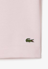 Tela de algodón de color rosa claro, con un pequeño logotipo de cocodrilo verde y blanco bordado cerca del dobladillo inferior. Textura suave, diseño clásico.