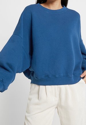 Femme portant un sweat-shirt bleu ample à manches longues et un pantalon en velours blanc taille haute, debout devant un fond uni.