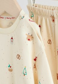Crème-kleurig loungekleding set bestaande uit een top en broek met feestelijke patronen van rendieren, de kerstman, bomen en pretzels. Zacht brei-materiaal.