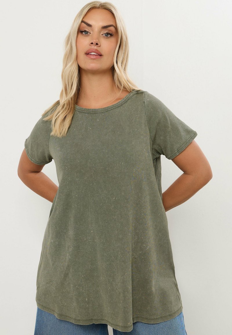 Donna bionda che indossa una maglietta verde oliva a maniche corte e larga e jeans blu, in piedi con le mani dietro la schiena.