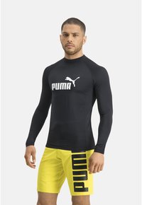 Schwarzes Rashguard mit langen Ärmeln und einem weißen Puma-Logo, kombiniert mit gelben Badehosen, die an der Seite einen großen schwarzen Puma-Druck zeigen.