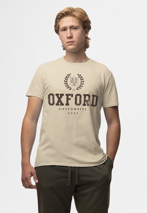 Jeune homme aux cheveux châtain clair, portant un t-shirt beige de l'université d'Oxford et un pantalon foncé, debout avec une main dans la poche.