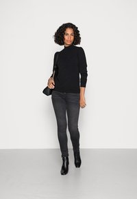 Maglione a collo alto nero in tessuto testurizzato, abbinato a jeans skinny grigio scuro e stivaletti neri con tacco. Piccola borsa nera a tracolla con catena.