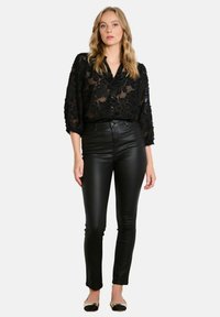 Zwarte doorschijnende blouse met bloemborduurwerk, gecombineerd met hoge zwarte nepleren broeken. Het model draagt zwarte platte schoenen.