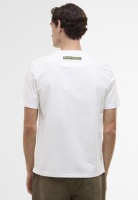 T-shirt branca de algodão com mangas curtas, apresentando um rótulo verde na parte de trás do pescoço. Design simples sem padrões ou texturas visíveis.